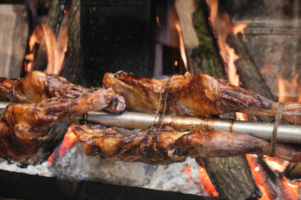 History of BBQ Sauce represented by roasting meat on a spit over an open fire
Alt Description: whole animal meat being roasted on a metal spit over an open outdoor fire for BBQ