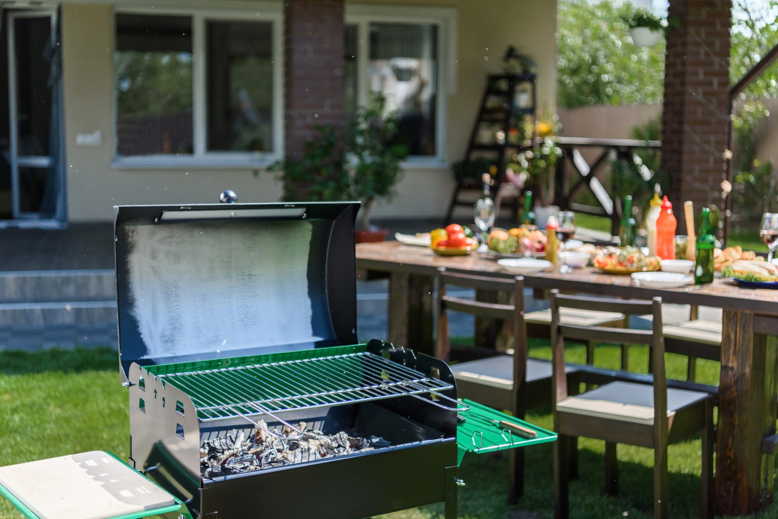 Outdoor grill with coals standing on green lawn and served table with dishes and drinks behind a backyard with a grill and table set up with BBQ food for a gathering